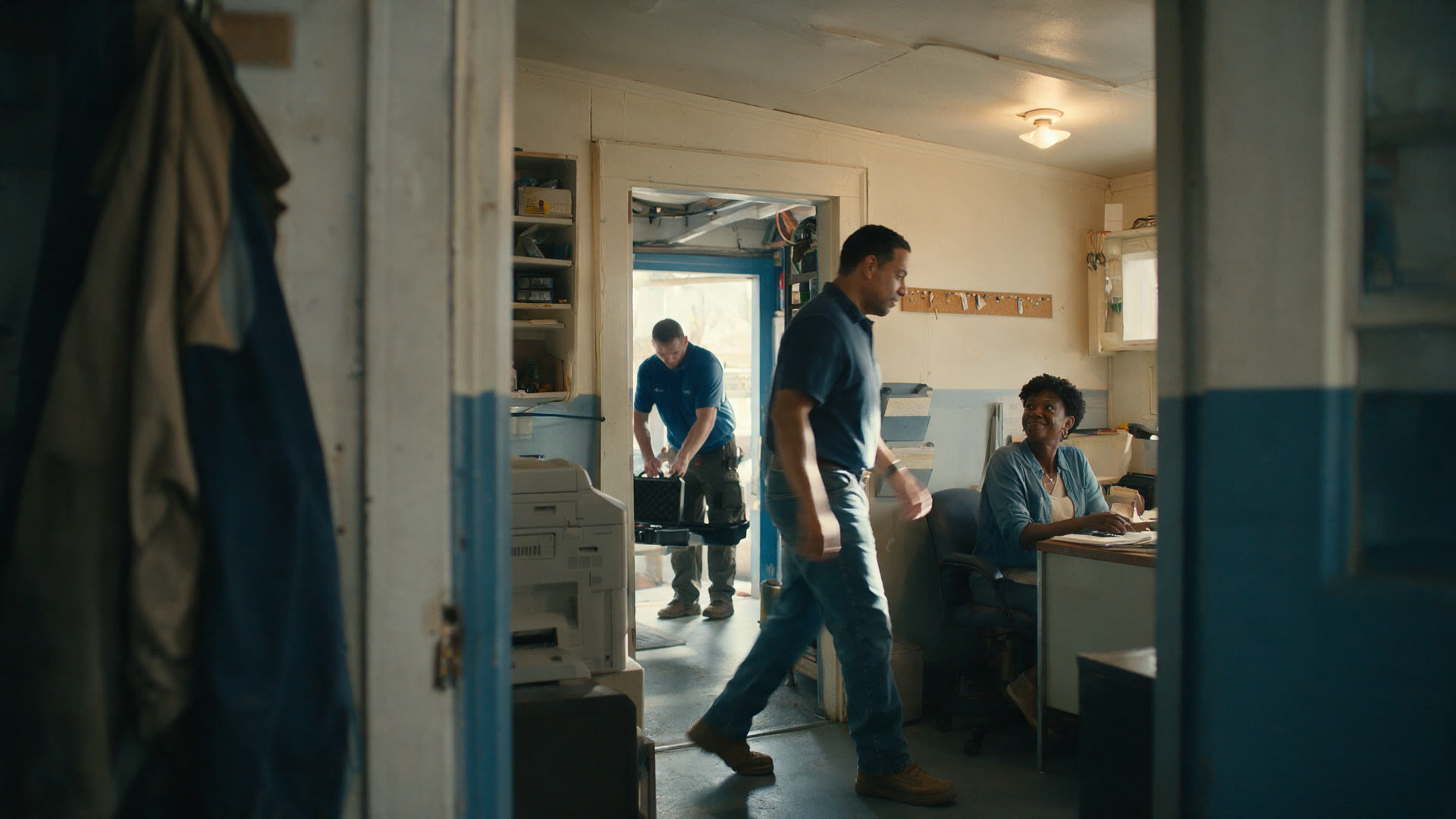 Two people walk through a small office past a desk where someone is working